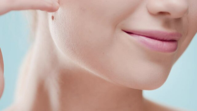 Extreme close-up of female beauty model touches her jawline smiling for the camera against blue background | Moisturizer wearing shot for face care commercial