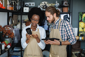 Happy african woman and caucasian man using digital tablet for receiving goods at decor store. Two colleagues in beige apron working together at modern shop.