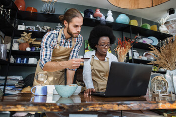 Two multicultural people in uniform analysing clients orders made on website of decor store. Handsome man holding credit card, charming woman typing on laptop.