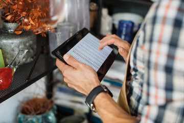 Cropped view from shoulder of caucasian seller in checkered shirt and apron using digital tablet for rechecking goods at decor store. Concept of sale, work and modern gadgets.