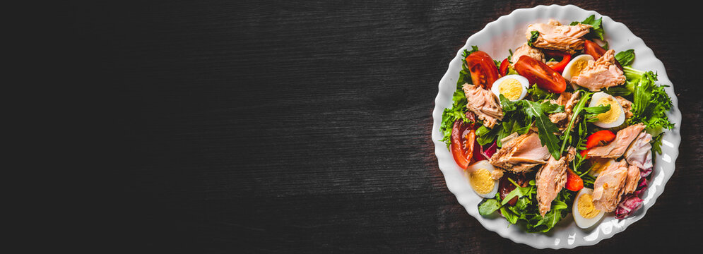 Fresh salad with fish, arugula, eggs,red pepper, lettuce, fresh sald leaves and tomato on a white plate on wooden table background