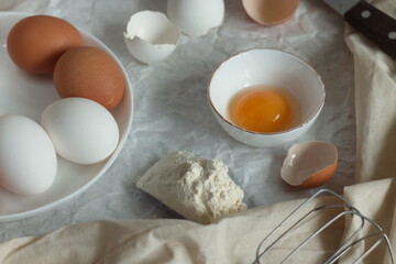 In the kitchen for cooking on the table - eggs on a plate, a broken egg with yolk in a bowl, a shell, a slide of flour and textiles