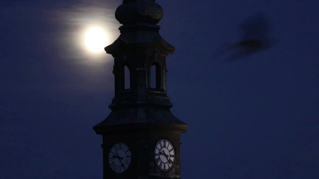 The full moon shines over the clock tower with flying birds in front.