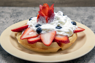 Decorative strawberry slices and sculpture stacked with blueberries and whipped cream on top of hearty waffle.
