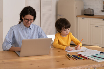 Young caucasian female work on laptop as son draw in notebook. Home office at covid-19 lockdown concept. Business woman mom busy with online management near calm kid. Mother entrepreneur at workplace