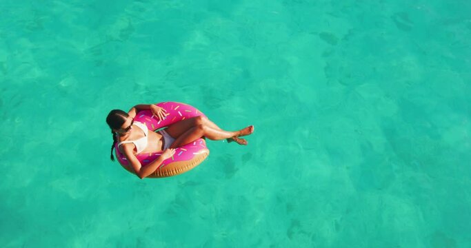 Beach vacation woman relaxing in pool float donut inflatable ring floating on turquoise ocean water background in Caribbean travel summer. Girl in white bikini top drone view.