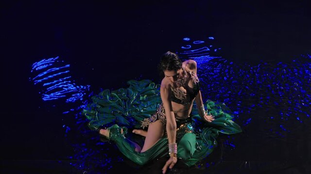 Top view of a wet oriental woman sitting on the surface of the water and dancing a belly dance. An exotic dancer with long black hair in a traditional costume performs in a dark studio. Slow motion.