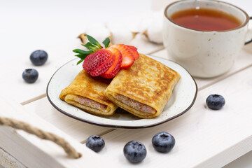 Russian rolled pancakes with strawberry filling. Served on a wooden tray with a cup of tea and berries.