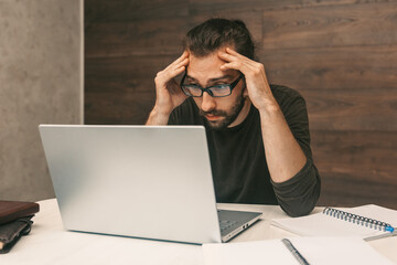 Work from home. The man works at home. Young man looking at laptop screen holding head with  hands