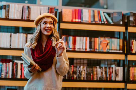 Portrait of girl, wearing sweater, hat and scarf, while looking at books from her favorite author at bookstore. Young blonde girls is book lover.