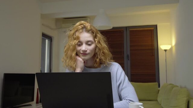 Cute girl sitting on a table in living room, infront of a laptop computer, drinking coffee. Portrait of a womam working from home. Camera moving left.