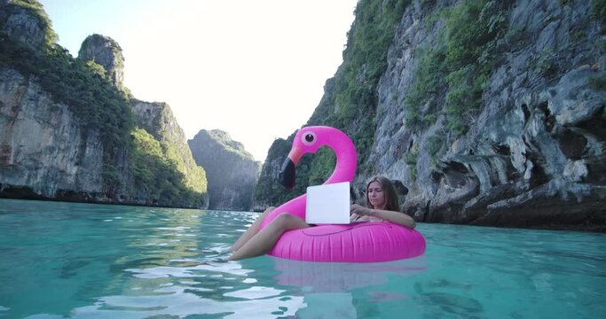 Young freelancer caucasian female floating on inflatable flamingo in the sea, remote working anywhere on her laptop computer connected to office and relax by seeing beautiful view on summer vacation.