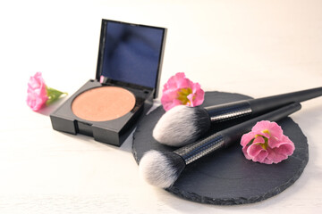 Obraz na płótnie Canvas Cosmetic brushes on a slate, make up face powder case and pink flowers on a white wooden background, beauty concept, copy space, selected focus