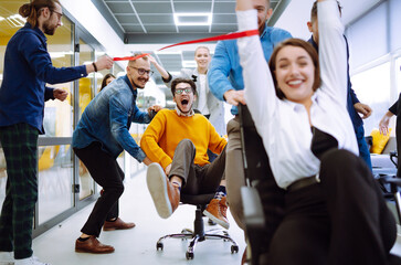 Friendly work team  ride chairs in office room cheerfully excited diverse employees laugh while enjoying fun work break activities, creative friendly workers play a game together. - obrazy, fototapety, plakaty