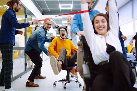 Friendly work team  ride chairs in office room cheerfully excited diverse employees laugh while enjoying fun work break activities, creative friendly workers play a game together.