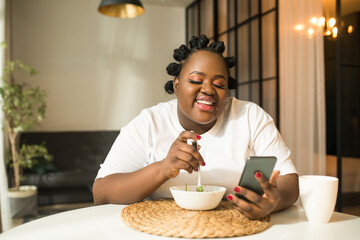 Fototapeta na wymiar Multiracial woman eating fresh salad with vegetables and using her smartphone
