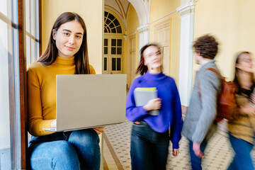 Brunette young woman student using laptop with interior of a busy university campus old building...