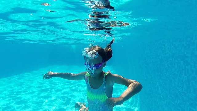 Pretty little 8s adorable girl wear swimsuit and goggles looking at camera enjoy summer vacations swimming in pool. Active lifestyle, summertime pleasure, sport activity concept. Slow motion view