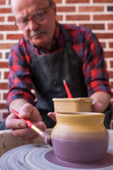 Fototapeta na wymiar Senior man holding a brush, painting on a ceramic using potter wheel.