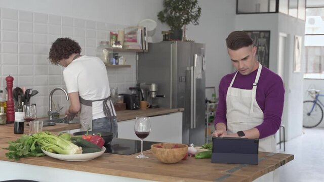 Lovely Young Gay Couple Enjoying Time Together In The Kitchen, Cooking Following A Recipe On Tablet, Drinking Wine And Washing The Dishes. Lgbt Daily Life Lifestyle Concept