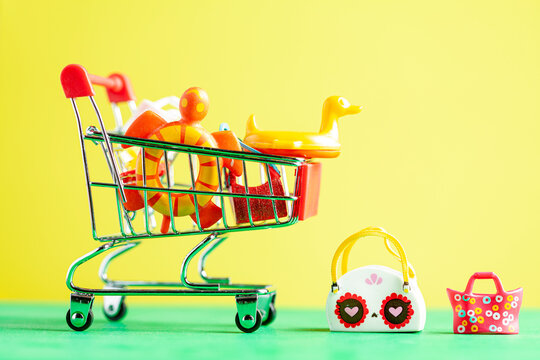 Abstract versatile image showing summer vacation shopping concept with beach bags and toys as well as duck floats are put inside the trolley with vibrant yellow background and green surface.