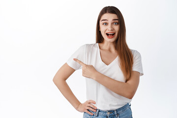 Hey take look. Excited beautiful girl smiling amazed, pointing finger aside at copyspace, showing advertisement, fantastic news, standing in t-shirt against white background