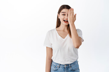 Surprised and happy girl covers half of face, smiling and gasping amazed, checking out something awesome, standing in casual clothes against white background