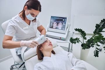 Charming Caucasian redheaded woman in white bathrobe receiving cleaning skin face with ultrasound...