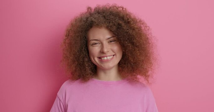 Positive good looking curly haired young woman with curly bushy hair waves hello smiles happily dressed in casual t shirt expresses good emotions isolated over pink background. Nice to meet you
