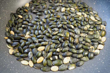 on top of the gray pan is a lot of green-colored roasted pumpkin seeds. side view . a gray frying pan on a dark table . cooking at home