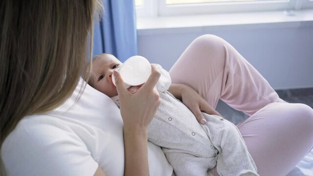 Long haired brunette bottle-feeds little naughty baby with pacifier sitting on bed under bright light against curtains and windows at home closeup