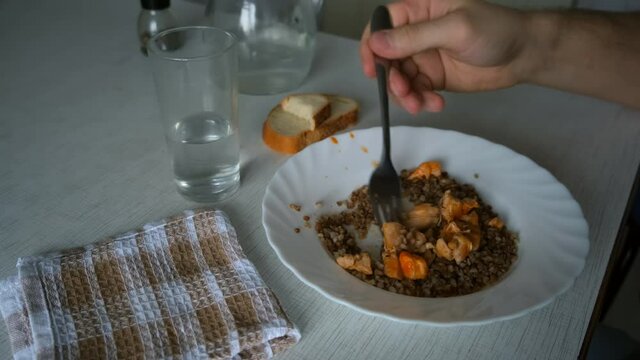 Man Is Eating Buckwheat And Chicken With Sauce And Bread, Drinking Water Sitting At Table At Home, Plate Closeup. Healthy Food Of Poor Person In Third World Country.