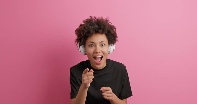 Upbeat cheerful dark skinned curly woman points index ingers at camera has fun dressed in casual clothes listens music via headphones expresses positive emotions isolated over pink background