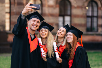group of happy students in bachelor's robes with diplomas taking a selfie on the phone outdoors.
