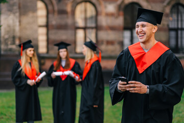 A cheerful, joyful male graduate, in a mantle, a cap, with a phone in his hand, is watching a funny video from the graduation ceremony.