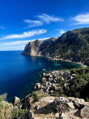 Playa de rocas con montañas llenas de vegetación un dia soleado con alguna nube en un mar calmado con un barco en verano