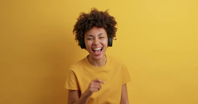 Overjoyed dark skinned woman laughs out points index finger at camera cannot stop laughing dressed in casual t shirt listens music from playlist via headphones isolated over vivid yellow wall