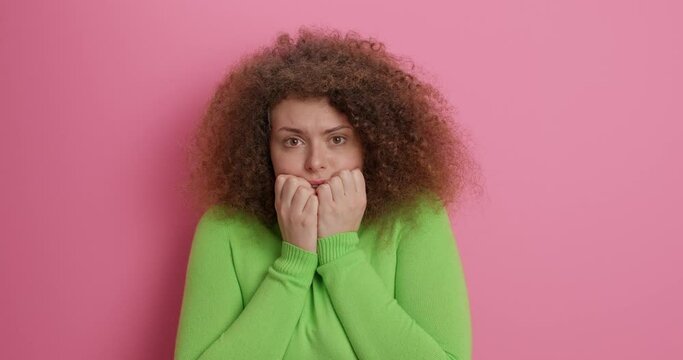 Scared beautiful woman with curly hair covers mouth and eyes looks with fright at camera has anxious scary look notices horrible scene wears casual green turtleneck isolated over pink studio wall