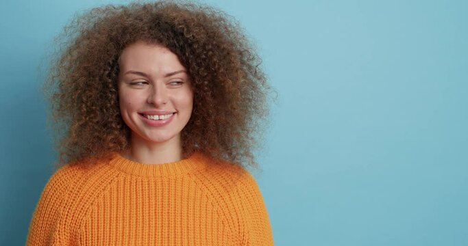 Pretty smiling woman has cutly hair has cheerful expression looks away gladfully pleased to hear good news wears casual knitted jumper isolated over blue background with copy space for text.