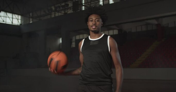 Video portrait of a basketball player bouncing a ball. African american man in uniform posing inside sports hall