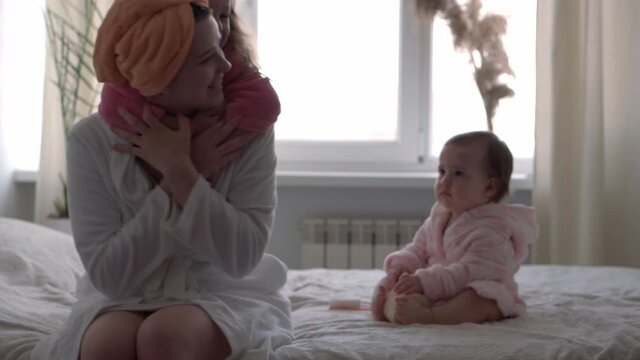 Authentic cute young woman hugging with baby girl. mom takes care of 2 children daughters different ages in bathrobes after bath on white bed oposite window. Spa, motherhood, childhood, family concept