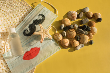  Protective disposable masks  with painted mustache and lips, beach hat, starfish, seashells and pebbles backlit by the sun on yellow background.  Summer 2021. The concept of travel during COVID-19