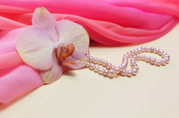 Peach phalaenopsis flower on pink chiffon drapery with pearl beads, composition with flower and jewelry, selective focus, horizontal orientation