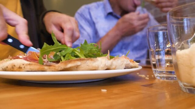 Time-lapse fast motion side view of adult and senior man - in background eating delicious pizza made from organic bio ingredients with fresh rucola arugula and prosciutto in traditional restaurant piz