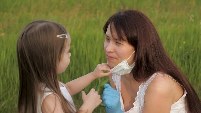 Mother wearing mask and gloves puts medical mask on child in park in summer on street during coronavirus epidemic. Mom and daughter in medical masks outdoors. Happy family mom, daughter hugging