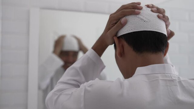 muslim asian man looking at mirror and get dressed before going to the mosque wearing islamic hat or cap