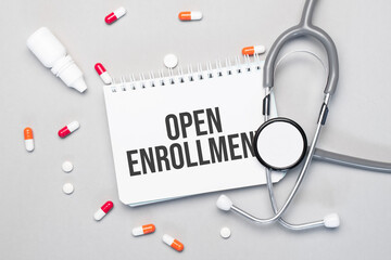 In the notebook is the text open enrollment next to a stethoscope, pills and glasses.