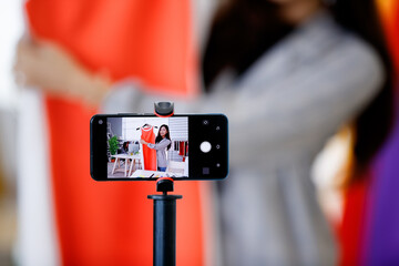 Closeup shot of black mobile smartphone set on tripod taking picture of Asian young pretty fashion designer in casual suit stand hold and show orange dress in hand while female colleague work at desk