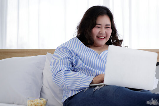 Asian young happy fat chubby woman lay down have fun laughing happily lean on white pillow on bed using laptop computer to watch movie and eat popcorn on quarantine lockdown on covid pandemic at home