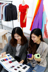 Fototapeta premium Two Asian young happy successful beautiful female creative stylish fashion designer colleagues hold fabric color pattern book and thread roll in hands talking discussing together on sofa in studio